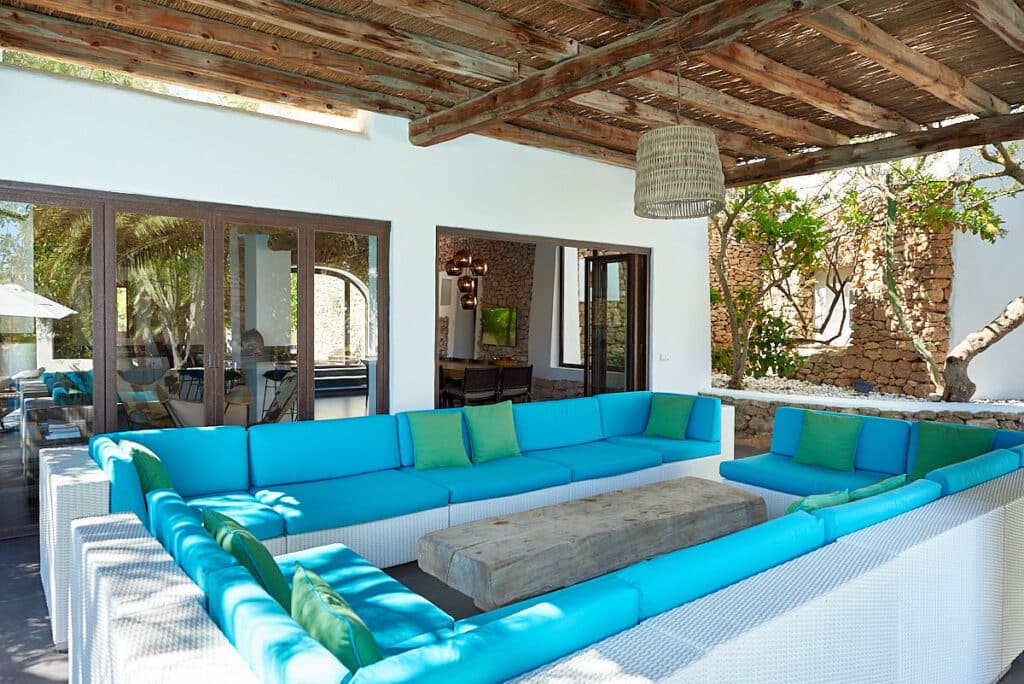 This outdoor patio exudes luxury travel vibes with a U-shaped white wicker sofa adorned with turquoise and green cushions. A rustic wooden coffee table commands the center, while overhead, a charming pergola sets the scene for relaxation next to a building featuring an open door and large windows. David Pearson Travel
