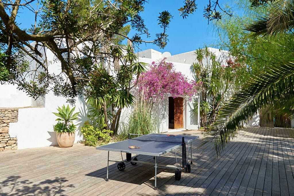 A ping pong table graces a spacious wooden deck, perfect for luxury travel enthusiasts. Surrounded by lush greenery and a white building, the scene is enhanced by trees, vibrant pink bougainvillea, and the serene backdrop of a clear blue sky. David Pearson Travel