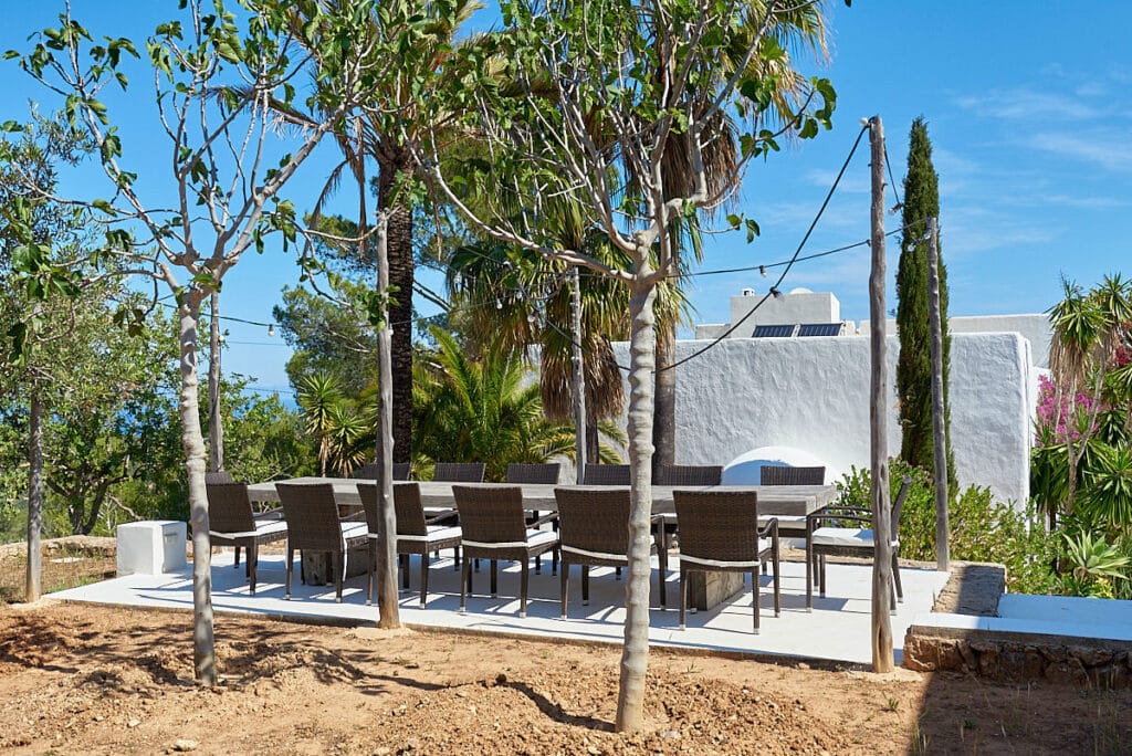 An outdoor dining area features a long table and several chairs under a canopy of trees, offering a touch of luxury travel. The garden setting is adorned with diverse plants and complemented by a white building in the background under a clear blue sky. David Pearson Travel