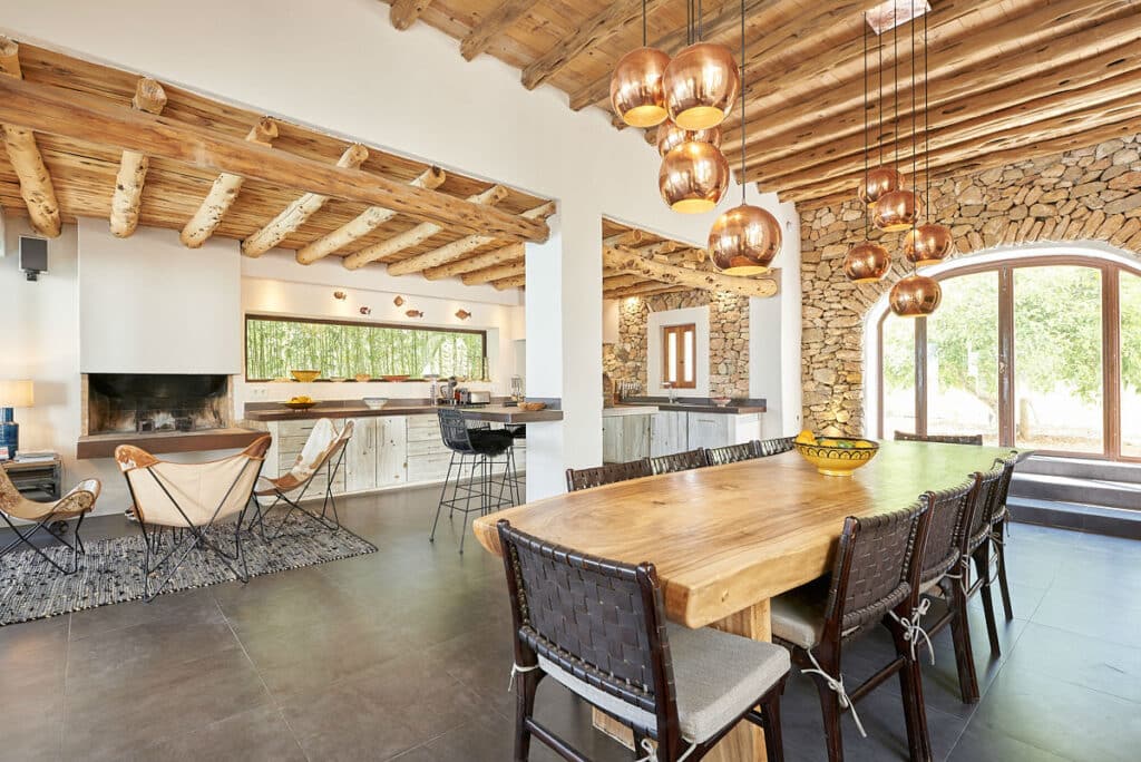 The spacious modern rustic kitchen and dining area evokes a sense of luxury travel with its wooden ceiling and large stone-framed windows. A wooden dining table surrounded by woven chairs sits beneath copper pendant lights. Natural light illuminates the space, enhancing the exquisite decor. David Pearson Travel