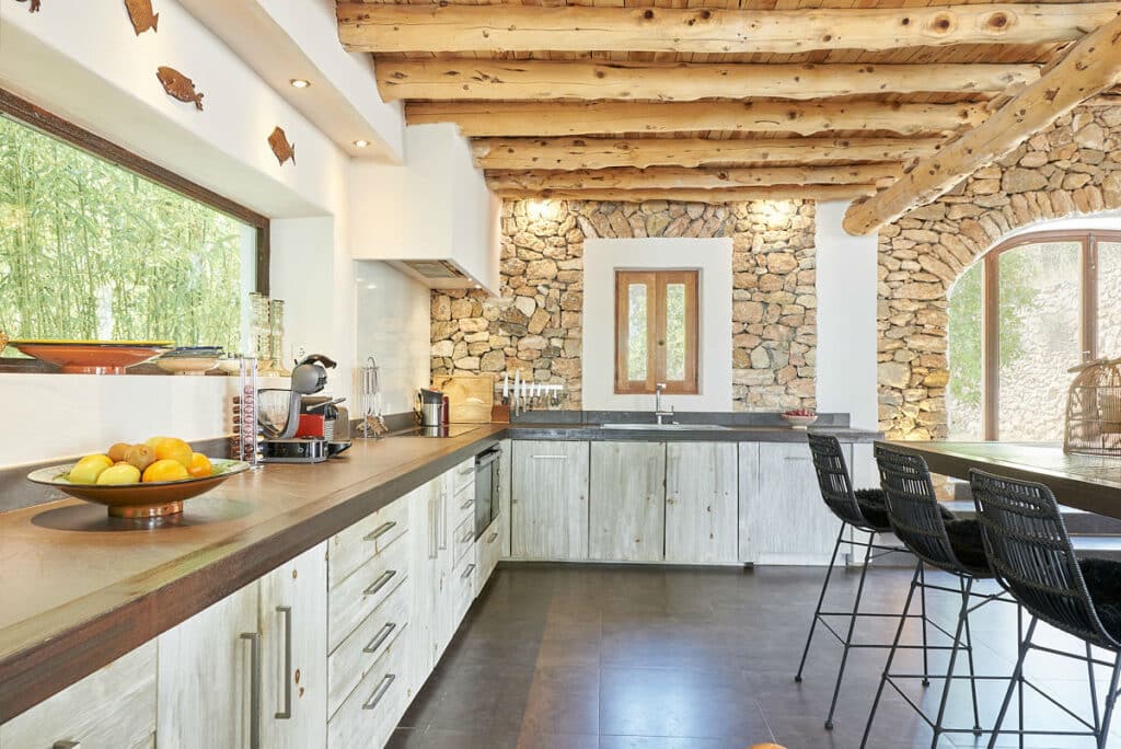Rustic kitchen with stone walls and wooden ceiling beams exuding the charm of a luxury travel retreat. Light wood cabinetry lines the counters, while a large window floods the space with natural light. Black stools surround a wooden island, accentuating a bowl of fruit and chic kitchen appliances. David Pearson Travel