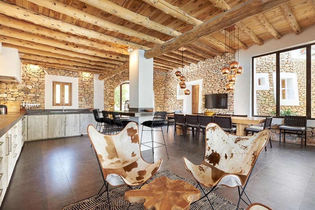 Rustic living area with wooden beamed ceiling, stone walls, and dark tile floor. It features butterfly chairs with cowhide upholstery, a wooden table, and modern pendant lights. Large windows offer natural light, creating an inviting space perfect for those seeking a touch of luxury travel-inspired decor. David Pearson Travel