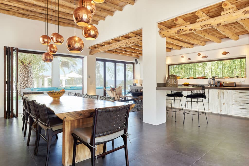 Spacious kitchen and dining area with rustic wooden ceiling and beams exudes a luxury travel vibe. Features a long wooden dining table, wicker chairs, modern pendant lights, open sliding glass doors, and a kitchen with white cabinets and stools at the counter. David Pearson Travel