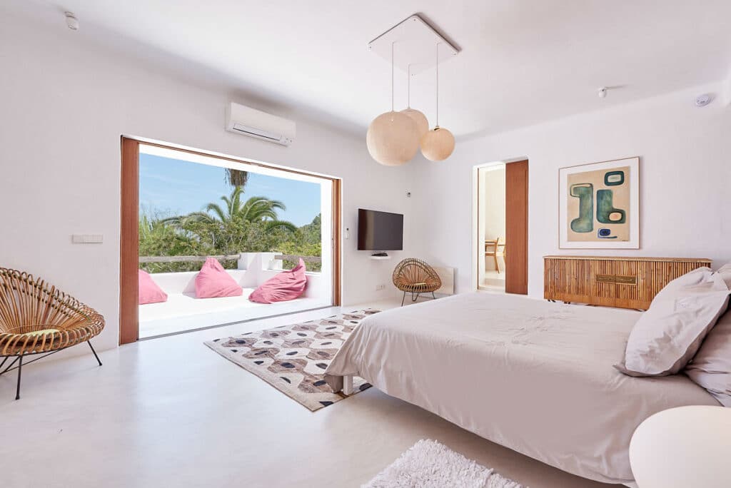 Modern bedroom with a large bed, wicker chairs, and a rug on a light floor. Glass doors open to a terrace with pink bean bags and a view of lush greenery. White walls are adorned with abstract art, globe lights hang from the ceiling, offering a luxury travel retreat atmosphere. David Pearson Travel