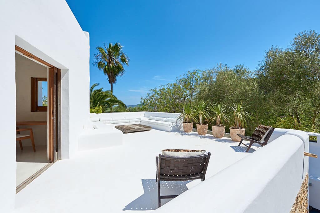 A sunlit terrace with white stucco walls, perfect for luxury travel, features cushioned seating and two wicker chairs. Potted plants line the railing, offering views of lush greenery and a palm tree under a bright blue sky. An open doorway invites you inside. David Pearson Travel