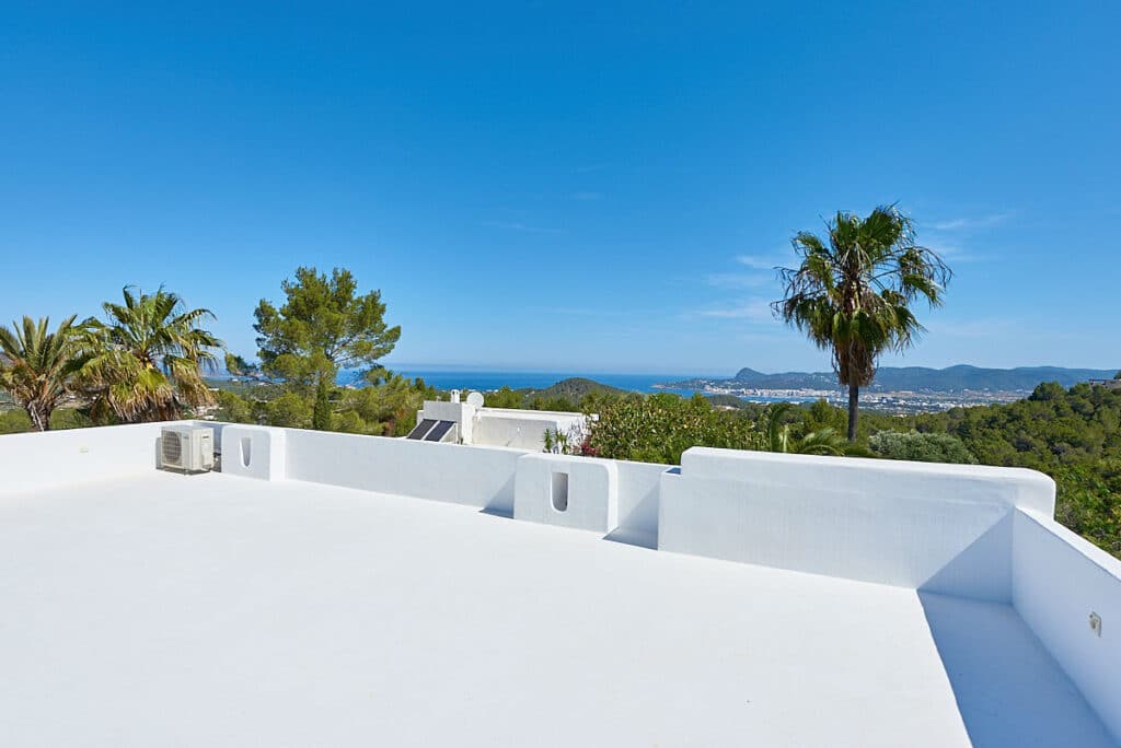 A pristine white rooftop terrace offers a luxury travel experience, overlooking lush greenery and palm trees with a distant view of the sea and mountains beneath a clear blue sky. David Pearson Travel