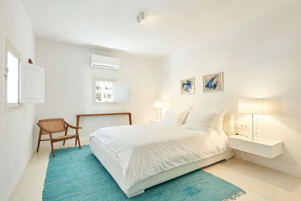 The minimalist bedroom exudes a luxury travel vibe with its white double bed, flanked by modern lamps on floating nightstands. A wooden chair and bench complement the serenity against the white wall, while a light blue rug adds color. Small artworks above the bed inspire wanderlust near the window. David Pearson Travel