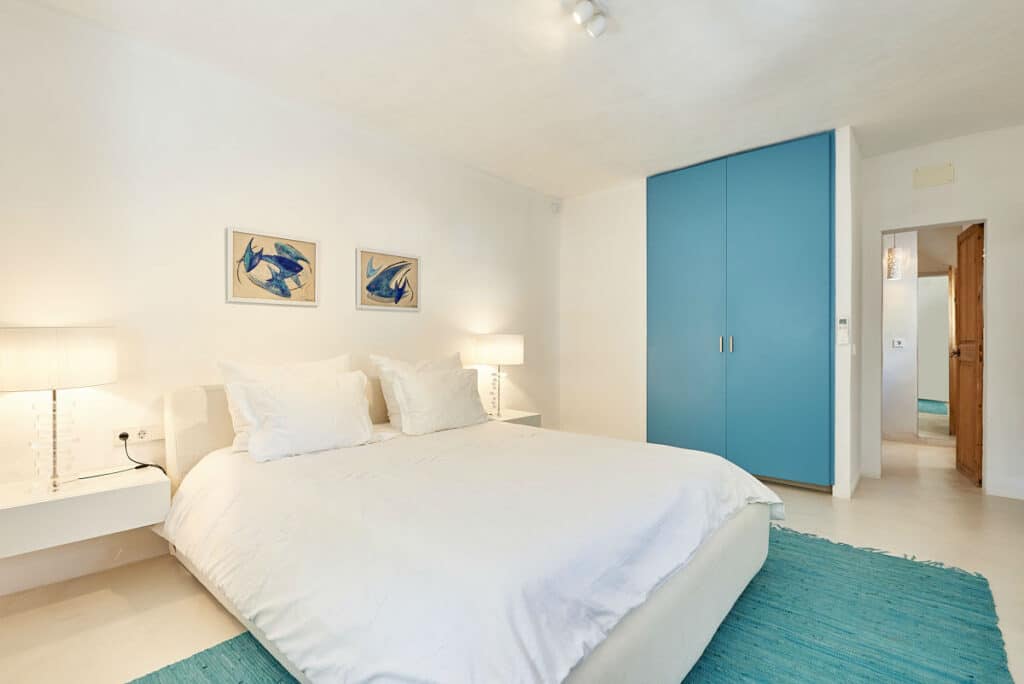 A bright bedroom exuding luxury features a large bed draped in white linens. Two elegant nightstands with lamps flank the bed, while sophisticated blue artwork adorns the wall above. A blue wardrobe and plush rug add a touch of opulence to this travel-inspired retreat. David Pearson Travel