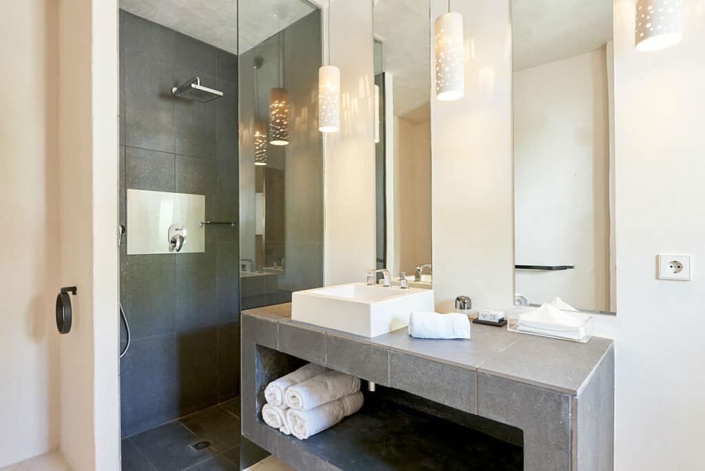 A modern bathroom exuding luxury travel vibes, with a glass-enclosed shower on the left featuring a dark tiled interior. On the right, a sleek vanity with a rectangular sink, folded towels, and mirror completes the look. Overhead, geometric pendant lights cast a warm glow. David Pearson Travel