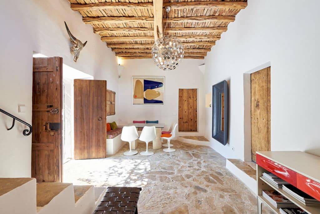Rustic interior with a stone floor and wooden beamed ceiling, perfect for luxury travel enthusiasts. Features a small dining area with four modern white chairs, colorful artwork, and natural wood doors. A large woven light fixture hangs overhead as sunlight streams through a side window. David Pearson Travel