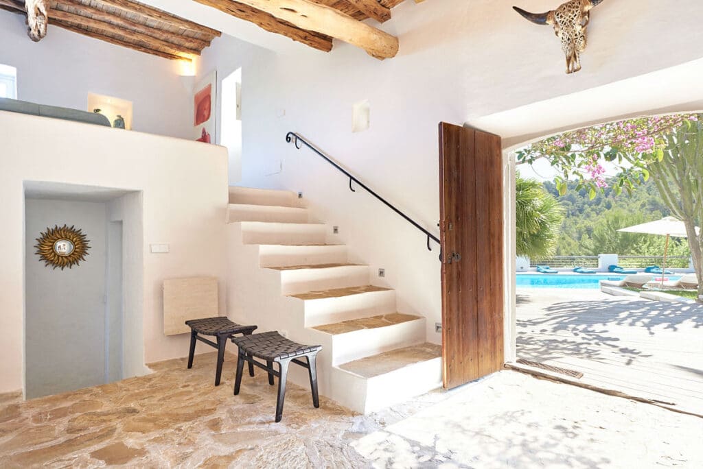 Bright Mediterranean-style interior with wooden beams and stone flooring exudes the charm of luxury travel. An open door reveals a pool and garden outside. Black chairs and stairs with a wrought iron railing enhance the cozy ambiance, while a decorative bull skull adorns the wall. David Pearson Travel