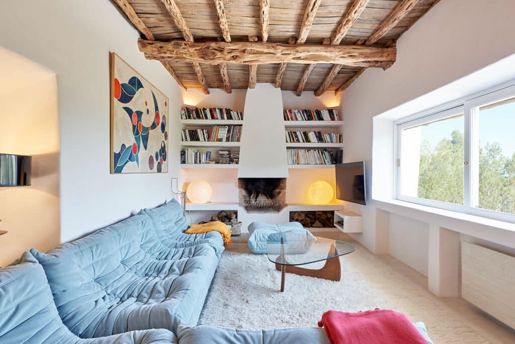 A cozy living room with a wooden beam ceiling exudes luxury travel vibes, featuring a blue sectional sofa, colorful wall art, and a fireplace flanked by bookshelves. A glass coffee table, large windows, and warm lighting create an inviting atmosphere reminiscent of an upscale retreat. David Pearson Travel