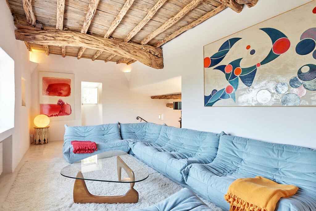 A stylish living room reminiscent of luxury travel, with a large blue sectional sofa accented by orange and yellow throws. It boasts a wooden beamed ceiling, abstract art on the wall, and a glass coffee table. Bright natural light filters through the windows. David Pearson Travel