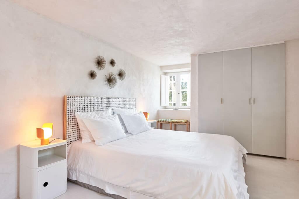 A minimalist bedroom with a touch of luxury travel features a white bed, plush pillows, and a checkered headboard. Soft lighting from bedside lamps on white tables enhances the ambiance. Three decorative wall pieces hang above the bed, with a large wardrobe and a window bench completing the serene retreat. David Pearson Travel