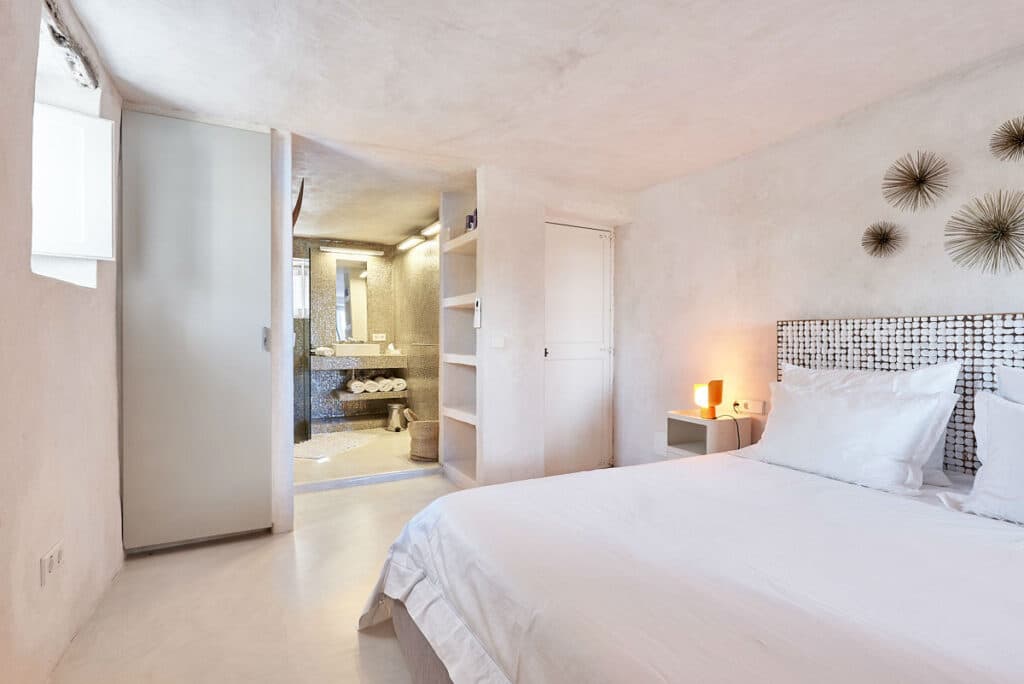 A minimalist bedroom featuring a white bed with a textured headboard and geometric wall decor, reminiscent of luxury travel destinations. Soft lighting from a nightstand lamp creates an inviting glow. An open door reveals a bathroom with neatly stacked towels and a decorative mirror. David Pearson Travel