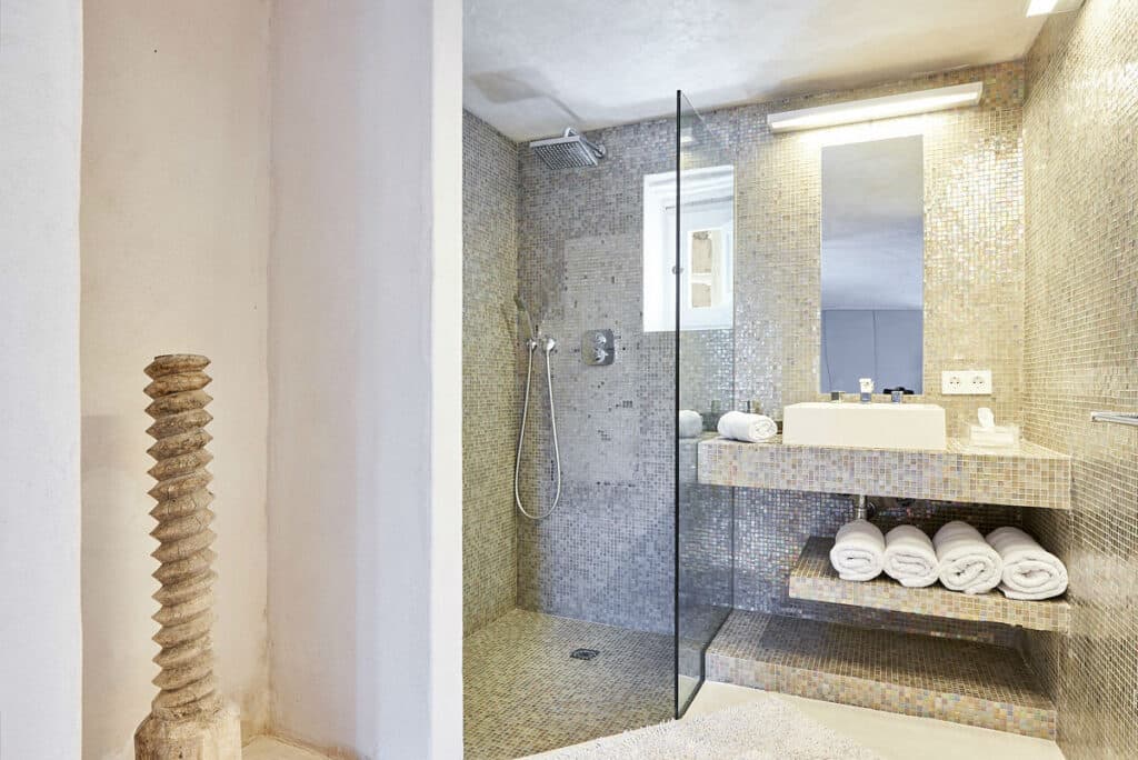 Modern bathroom with a glass-enclosed shower adorned in mosaic tiles, evoking luxury travel experiences. A rectangular mirror sits above a white sink with stacked towels below. On the left, a decorative, textured column stands against a pristine white wall, exuding sophistication. David Pearson Travel