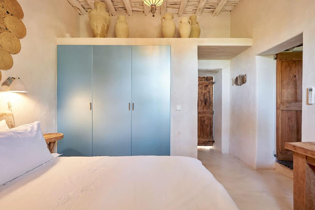 A bedroom with cozy, rustic charm perfect for luxury travel enthusiasts. It features a wooden bed adorned with white bedding, blue wardrobes, and wicker baskets on the wall. The wooden doors and exposed beams enhance its inviting ambiance. David Pearson Travel