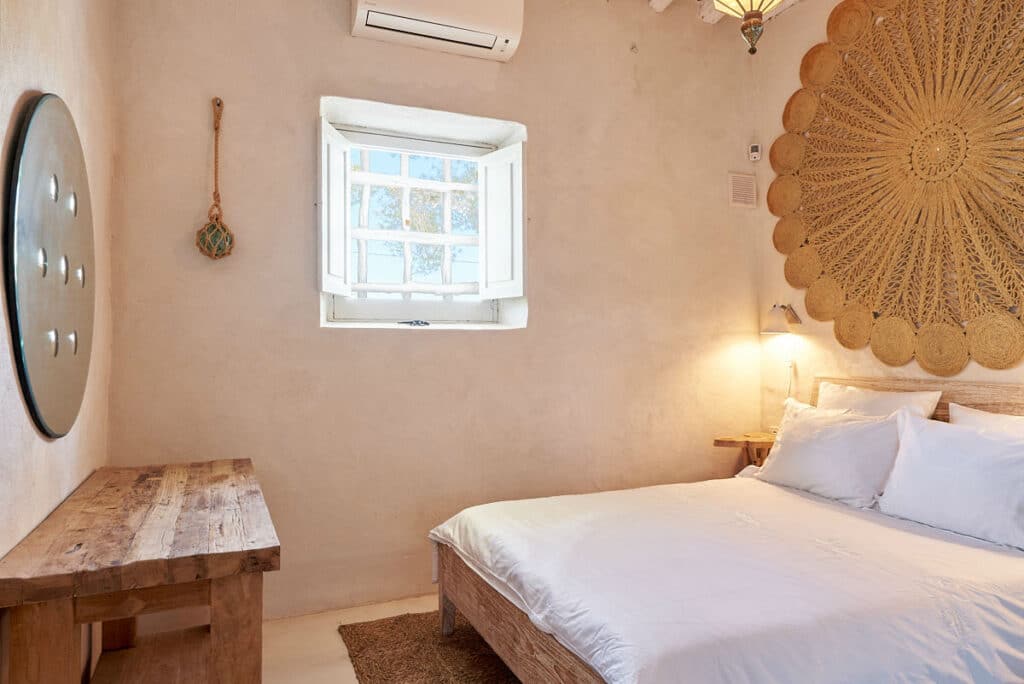 Cozy bedroom with a rustic wooden bed and bedside table, complemented by a round wall decoration and a small window letting in natural light. Beige walls, an air conditioner, and a hanging rope ornament create an inviting retreat for your luxury travel experience. David Pearson Travel