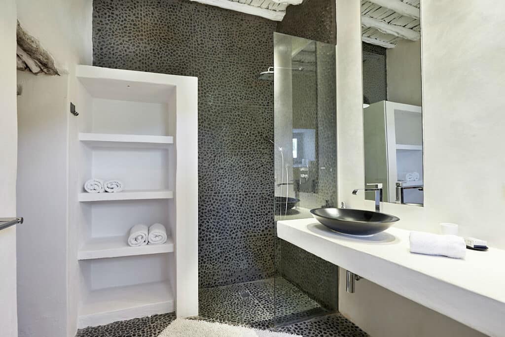 The modern bathroom exudes the ambiance of luxury travel, with white walls and a pebble-textured shower area. It features open shelves with rolled white towels, a sleek dark basin on a white countertop, and a glass shower partition. David Pearson Travel