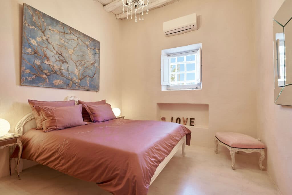 A cozy bedroom exudes the charm of luxury travel with a bed adorned in pink bedding, a chandelier, and a painting of branches on the wall. A window with an air conditioner graces the decorative LOVE display. A small cushioned bench sits to the right, while soft lighting creates a warm ambiance. David Pearson Travel
