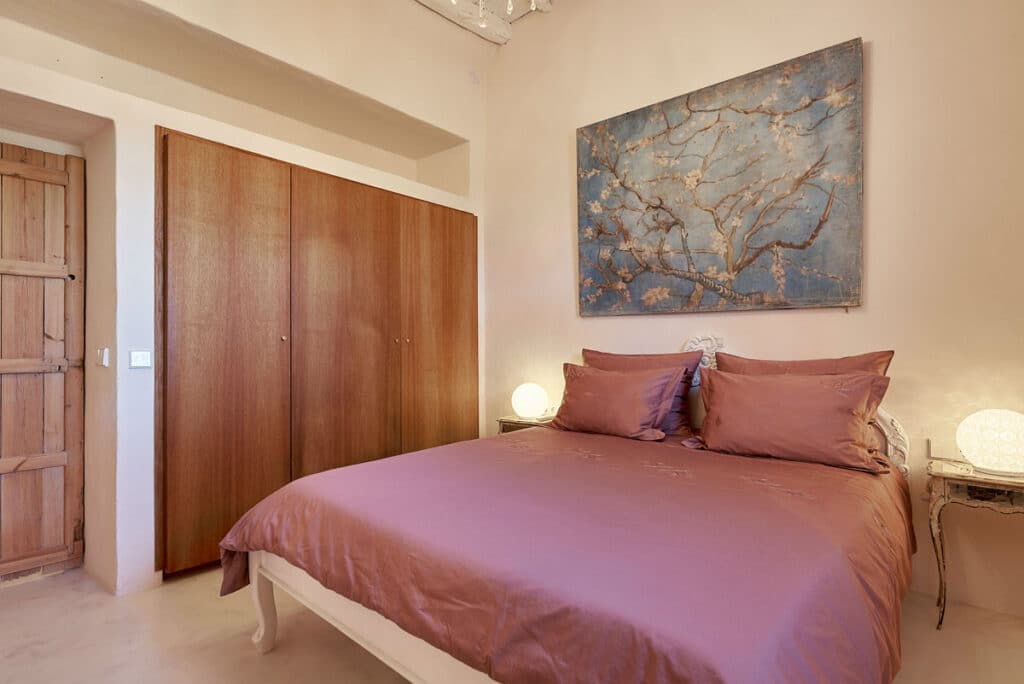 A cozy bedroom with a mauve bedspread and matching pillows epitomizes luxury travel. Two bedside tables with spherical lamps flank the bed, casting a warm glow. Above, a large painting of tree branches on a blue background adds elegance. A wooden wardrobe and door complete this tranquil haven. David Pearson Travel