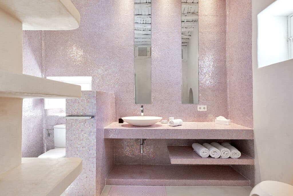 Elegant bathroom with light pink mosaic tiles, featuring a modern white sink and faucet reminiscent of luxury travel. Two tall mirrors are mounted above, and white rolled towels are neatly arranged on a shelf beneath the counter. David Pearson Travel