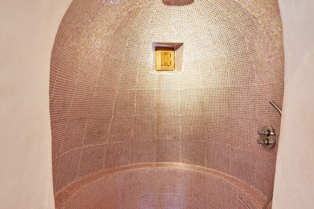A rounded, mosaic-tiled shower with pinkish tiles curves along the wall and ceiling, exuding the ambiance of luxury travel. It features a small, square window and chrome shower controls on the right side, offering a cozy, enclosed feel. David Pearson Travel