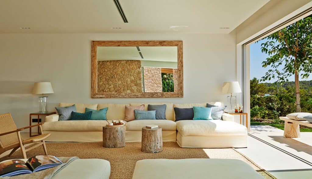 A spacious living room with a large white sofa adorned with colorful cushions evokes the ambiance of luxury travel. A wooden-framed mirror hangs on the wall, while two tree stump side tables hold accessories. Natural light streams through a large glass door, revealing lush greenery outside. David Pearson Travel