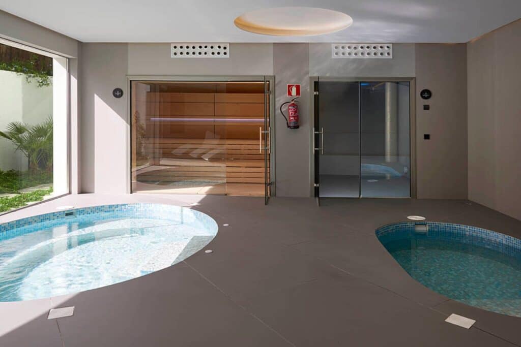 This luxury travel oasis features an indoor spa with two circular hot tubs in a tiled room. A glass-enclosed sauna and steam room with wooden interiors are in the background. Sunlight pours through a window, casting a warm glow next to the mounted fire extinguisher. David Pearson Travel