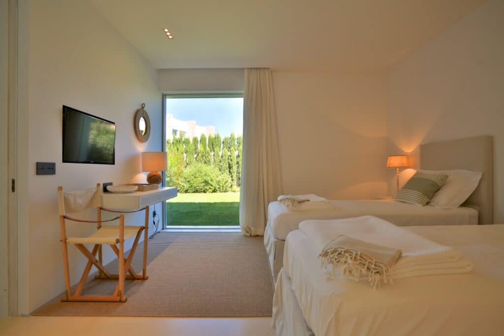 A minimalist bedroom designed for luxury travel, featuring two single beds with white bedding and light beige decor. A large window reveals a lush green garden. The room includes amenities like a wall-mounted TV, a small desk with chair, and a lamp on the bedside table. David Pearson Travel