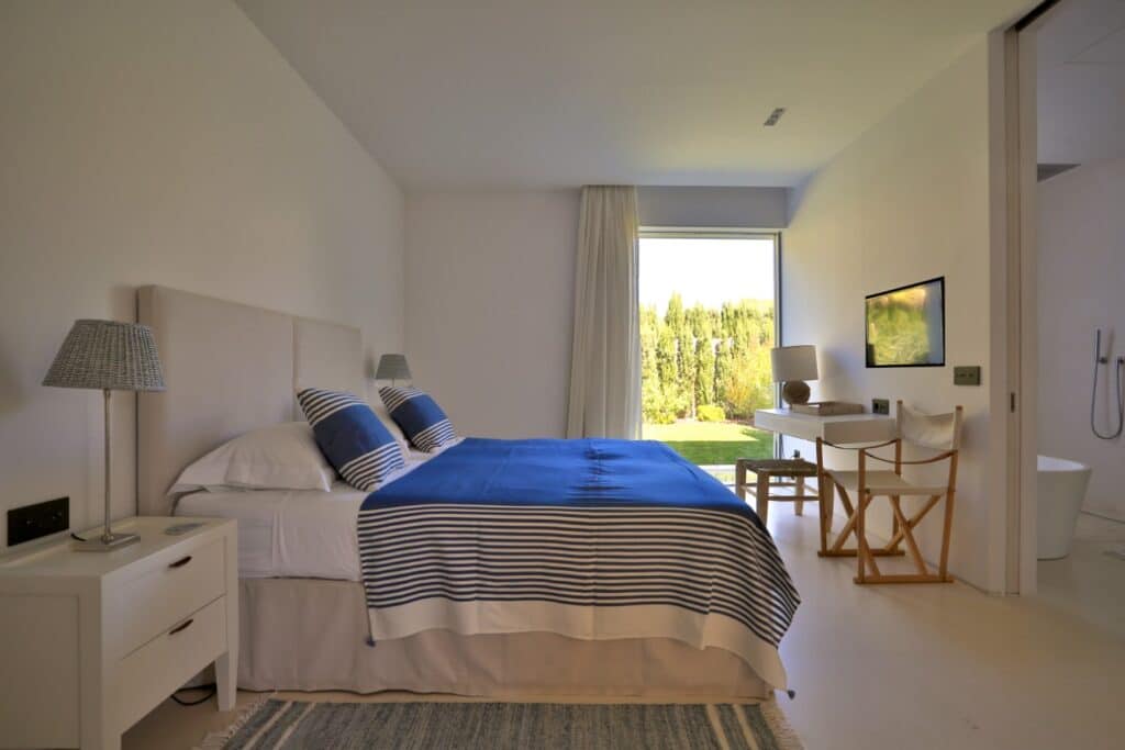 Modern bedroom with a large double bed featuring blue and white bedding, embodying a luxury travel vibe. A window offers views of greenery outside. The room includes pristine white walls, a plush rug, two bedside tables with elegant lamps, a desk, and a chair. David Pearson Travel