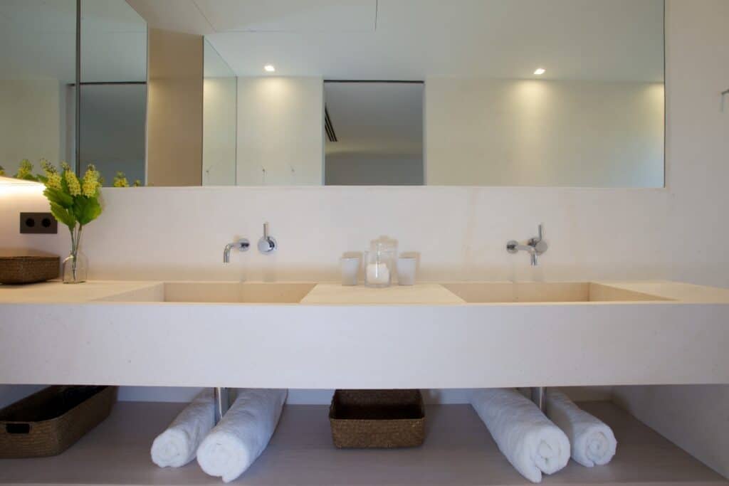 A modern bathroom exuding luxury travel vibes features a large rectangular mirror above a double sink vanity. Two rolled towels are neatly placed in baskets under the sinks, while a small vase with flowers and toiletries adorns the countertop, adding an elegant touch. David Pearson Travel