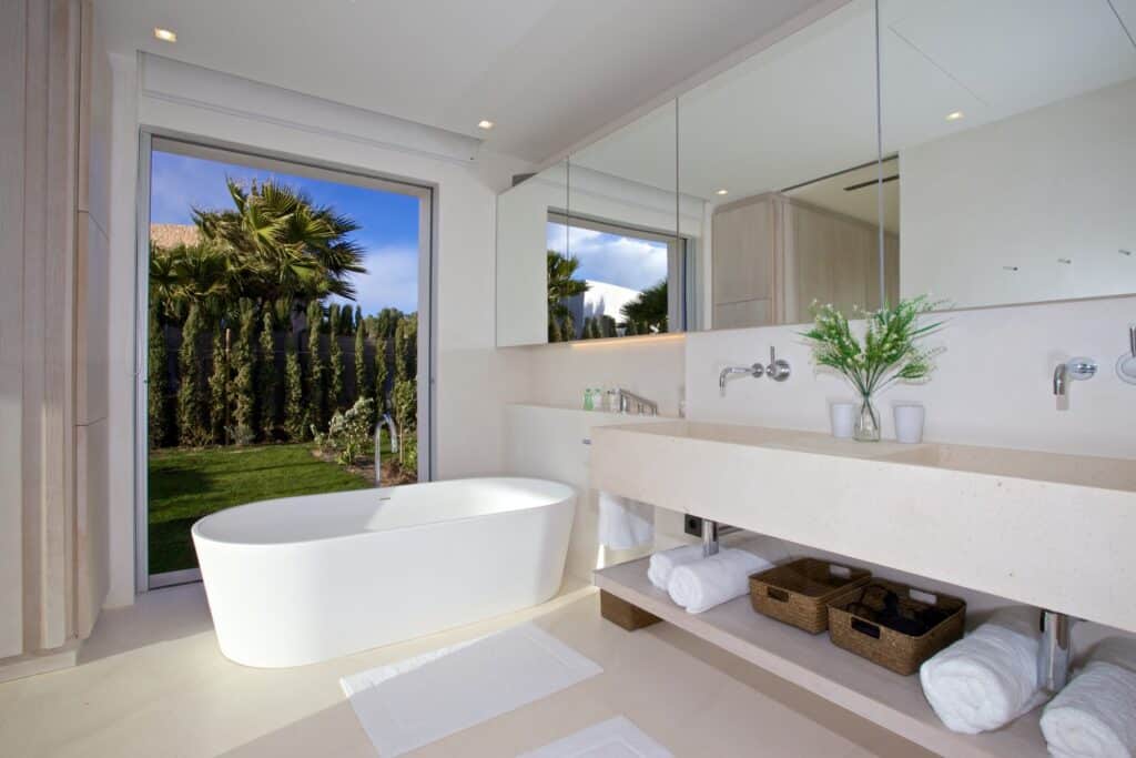 Luxurious bathroom blending the essence of luxury travel, featuring a freestanding white bathtub by a window with garden views. A beige double sink vanity, large mirror, and storage baskets complete the serene space in neutral tones and modern design. David Pearson Travel
