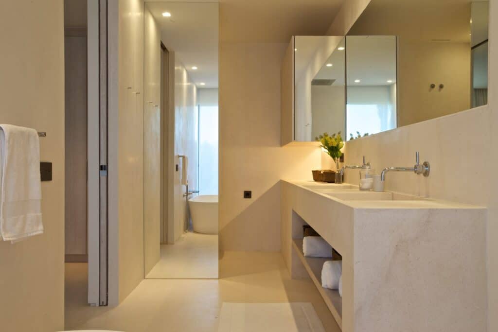 Modern bathroom with beige walls and flooring epitomizes luxury travel-inspired design. Features a large mirror above a double sink with a countertop. Towels are neatly stacked below, and a vase with yellow flowers sits on the counter. A freestanding bathtub graces the background. David Pearson Travel