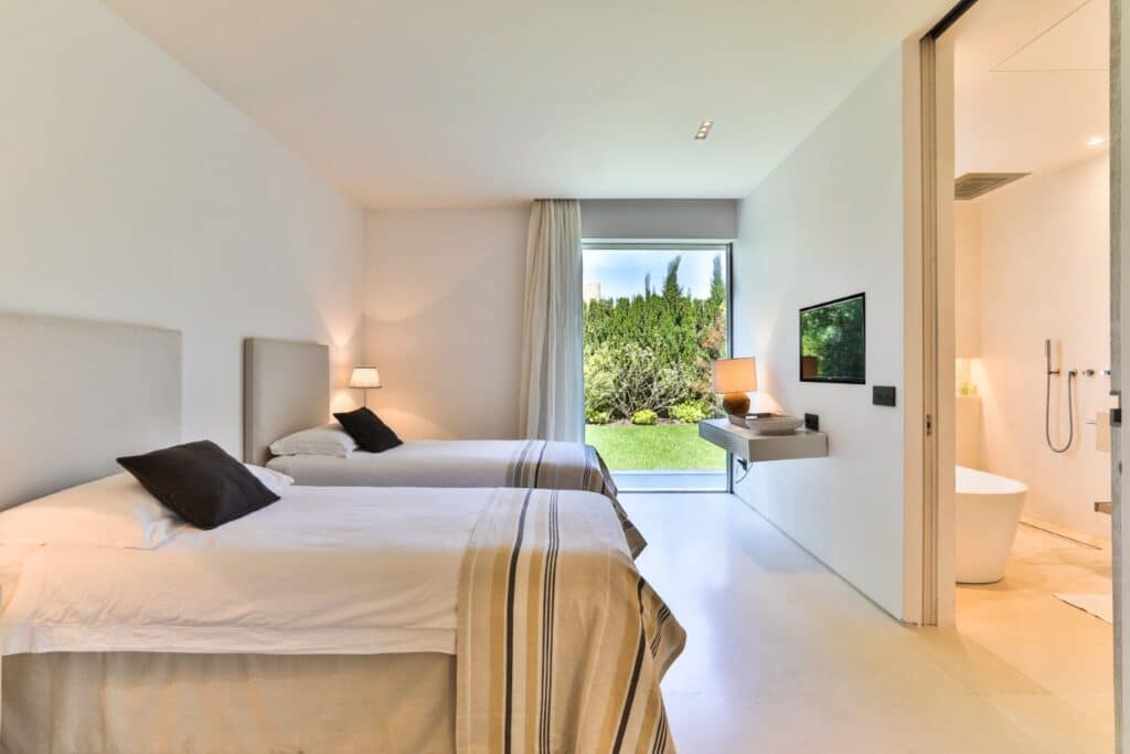Minimalist bedroom with two beds showcasing striped bedding ideal for luxury travel. A desk with a lamp sits beneath a wall-mounted TV. Large window offers a lush garden view. Adjacent bathroom visible, featuring a walk-in shower and freestanding bathtub in a neutral color palette. David Pearson Travel