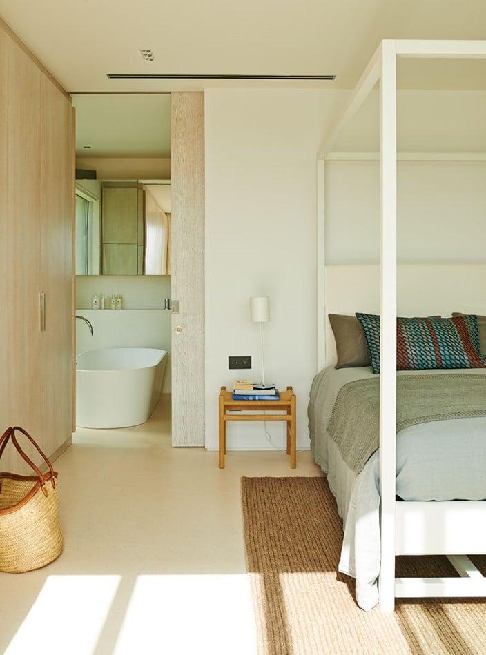 Minimalist bedroom featuring a four-poster bed, grey bedding, and colorful pillows. A woven rug and basket adorn the floor. A small wooden stool stacked with books foreshadows adventures in luxury travel. A sliding door opens to a bathroom with a modern freestanding tub. David Pearson Travel