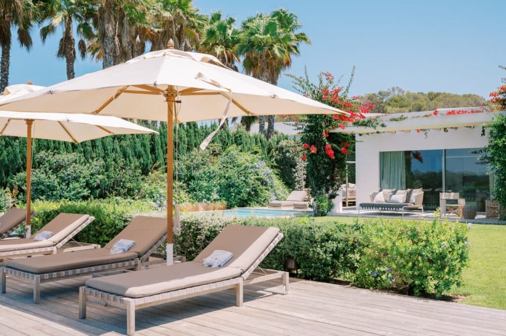 Lounge chairs with white umbrellas are set on a wooden deck beside a lush green garden, perfect for luxury travel relaxation. A modern house with large windows stands in the background, surrounded by trees and colorful flowers under a clear blue sky. David Pearson Travel