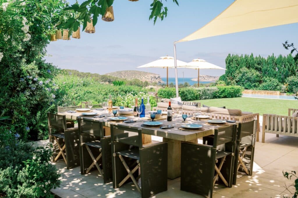 The outdoor dining area, ideal for luxury travel enthusiasts, features a large wooden table set for a meal and surrounded by green chairs. Overhead, a canvas sunshade and hanging lanterns provide shade. In the background, lush greenery frames the pool with distant views of hills and sea. David Pearson Travel