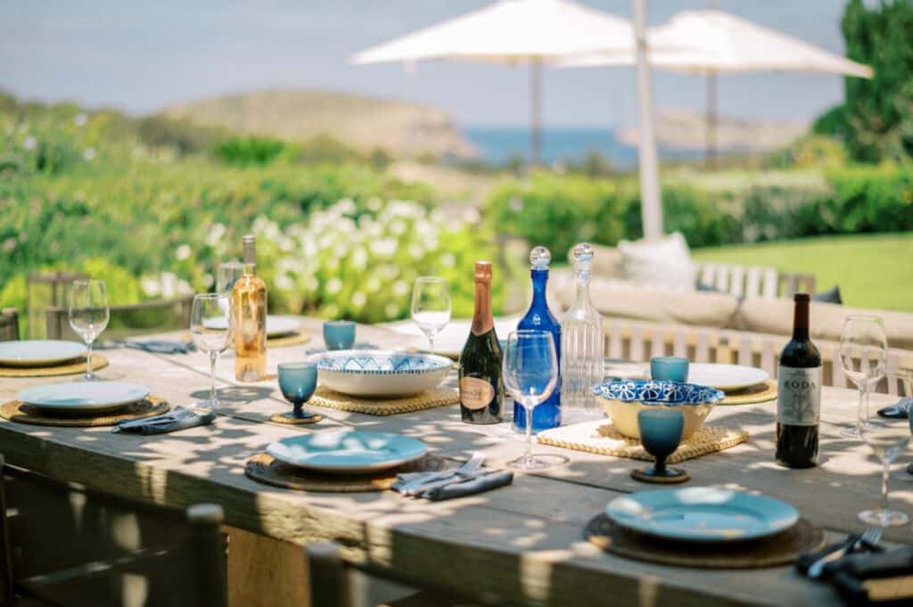 A luxurious wooden outdoor table is set for a meal, featuring plates, glasses, and bottles of wine. Blue patterned bowls add charm amid sunny, lush greenery. Large white umbrellas offer shade while the breathtaking sea view completes this luxury travel experience. David Pearson Travel