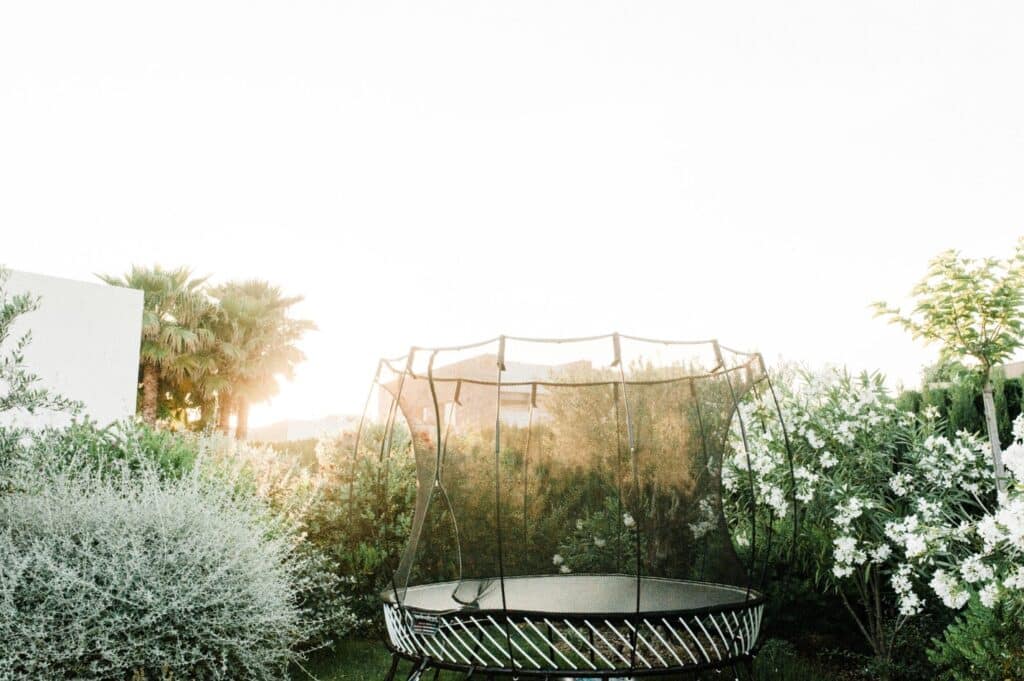A backyard scene with a black trampoline surrounded by lush green plants and bushes exudes a sense of luxury travel. The setting sun casts a warm glow, reminiscent of an exotic getaway, while palms and white flowering plants complete the serene vista. David Pearson Travel