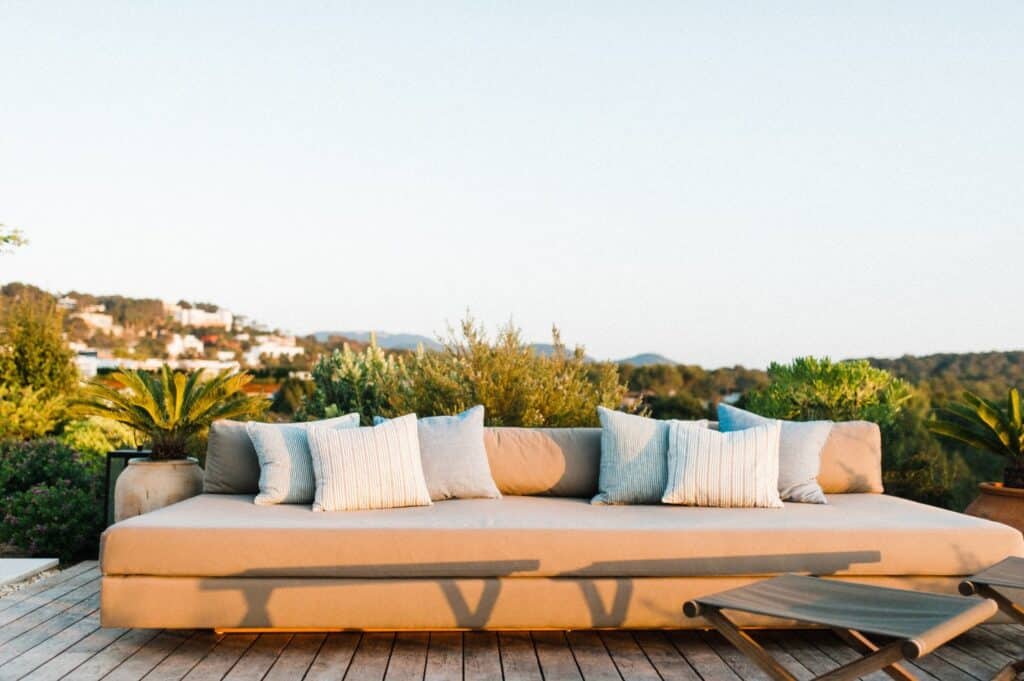 A modern outdoor patio boasts a large beige sofa with white and gray cushions, perfect for luxury travel relaxation. It overlooks scenic hills and greenery under a clear blue sky, with a sun lounger inviting you to unwind on the wooden deck. David Pearson Travel