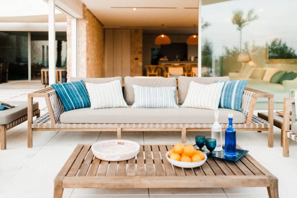 A modern outdoor patio exudes a luxury travel vibe with a wooden couch adorned with blue and white striped cushions. A wooden coffee table displays a bowl of oranges, a tray with bottles, and glasses, while glass doors reveal an indoor dining area ready for exquisite experiences. David Pearson Travel