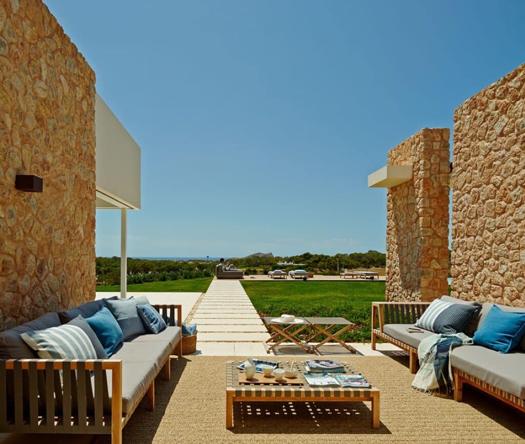 The outdoor patio exudes luxury travel vibes with its stone walls and two wooden sofas adorned with blue and white cushions. A large rug anchors the coffee table at the center, while a lush green lawn unfolds towards a scenic view under a clear blue sky. David Pearson Travel