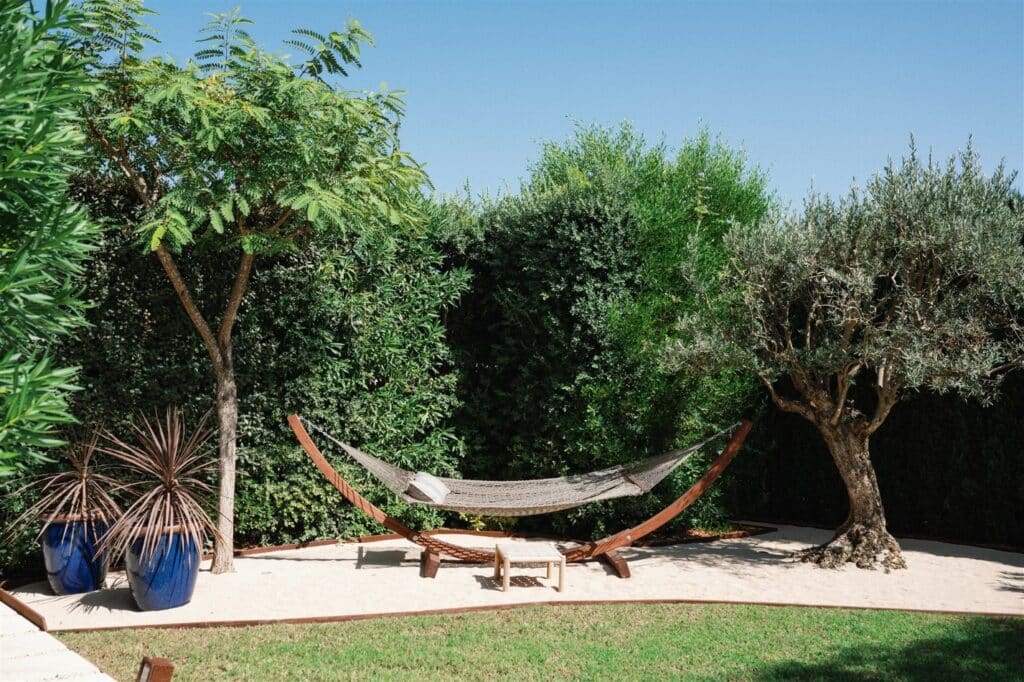 A serene backyard scene with a hammock stretched between two trees, surrounded by lush greenery, evokes the tranquility of luxury travel. Two plants in blue pots sit on the left, while a small wooden stool waits by the hammock beneath a luxurious, clear blue sky. David Pearson Travel