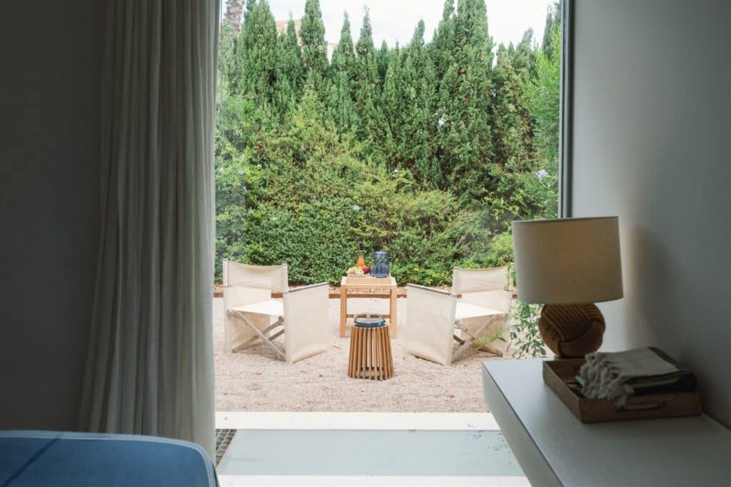 From the room, one can appreciate a patio with four beige chairs encircling a small table. This serene oasis, embraced by lush greenery, invites moments of quiet luxury. In the foreground, a lamp and book hint at indulgent travel reflections in this tranquil retreat. David Pearson Travel
