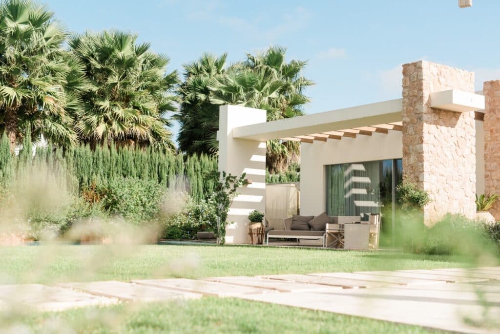 Modern house with stone and white walls, large windows, and a patio with seating under a pergola. Perfect for luxury travel, its manicured lawns, palm trees, and lush bushes create a serene outdoor space. David Pearson Travel