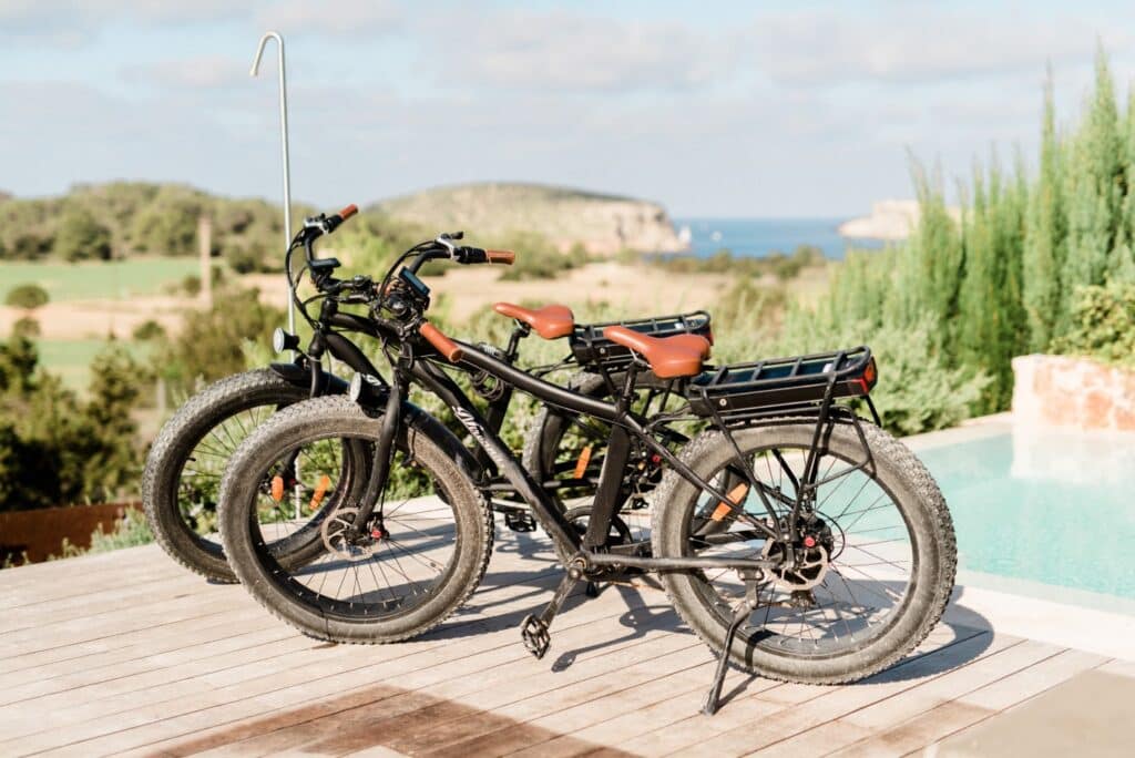Three electric bikes with wide tires and brown seats are parked on a wooden patio, epitomizing luxury travel. The background boasts a scenic view of greenery, a distant coastline, and a clear blue sky. David Pearson Travel