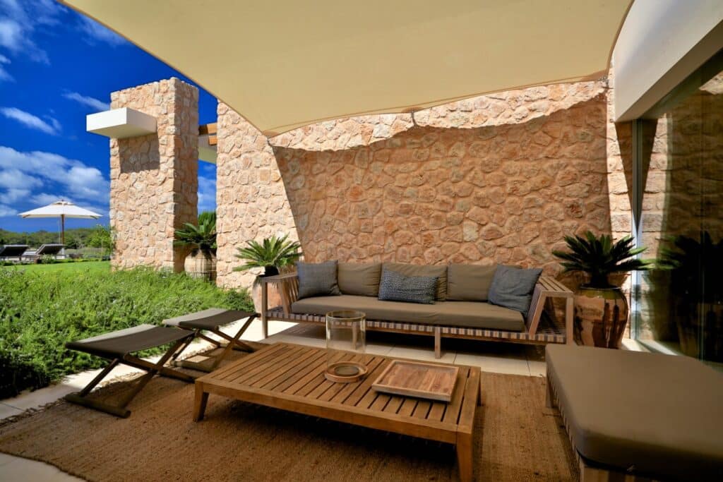 The patio area exudes the essence of luxury travel, boasting a stone wall backdrop, a tan outdoor sofa with gray cushions, and a wooden coffee table. It features two lounge chairs on a woven rug under a large white canopy, surrounded by lush greenery and set against a clear blue sky. David Pearson Travel