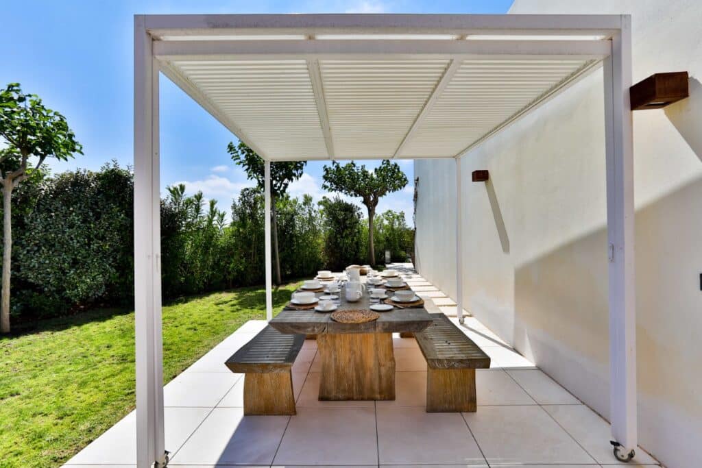 The outdoor dining area exudes luxury travel vibes, featuring a long wooden table adorned with elegant plates under a modern white pergola. Surrounded by lush green bushes and a sunny sky, the space boasts wooden benches and rests gracefully on a tiled patio adjacent to a pristine white wall. David Pearson Travel