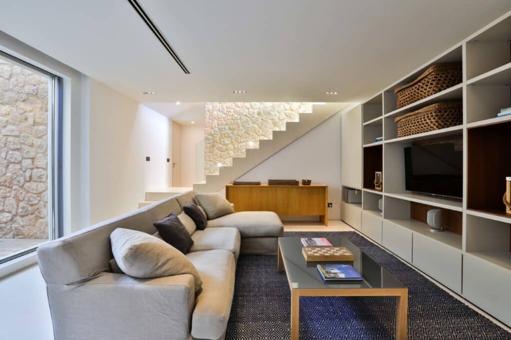 A modern living room features a light gray sectional sofa and a glass coffee table with books, evoking a sense of luxury travel. Built-in shelves adorned with baskets and decor, along with a stone-textured wall by the stairs and large window, complete the cozy ambiance. David Pearson Travel